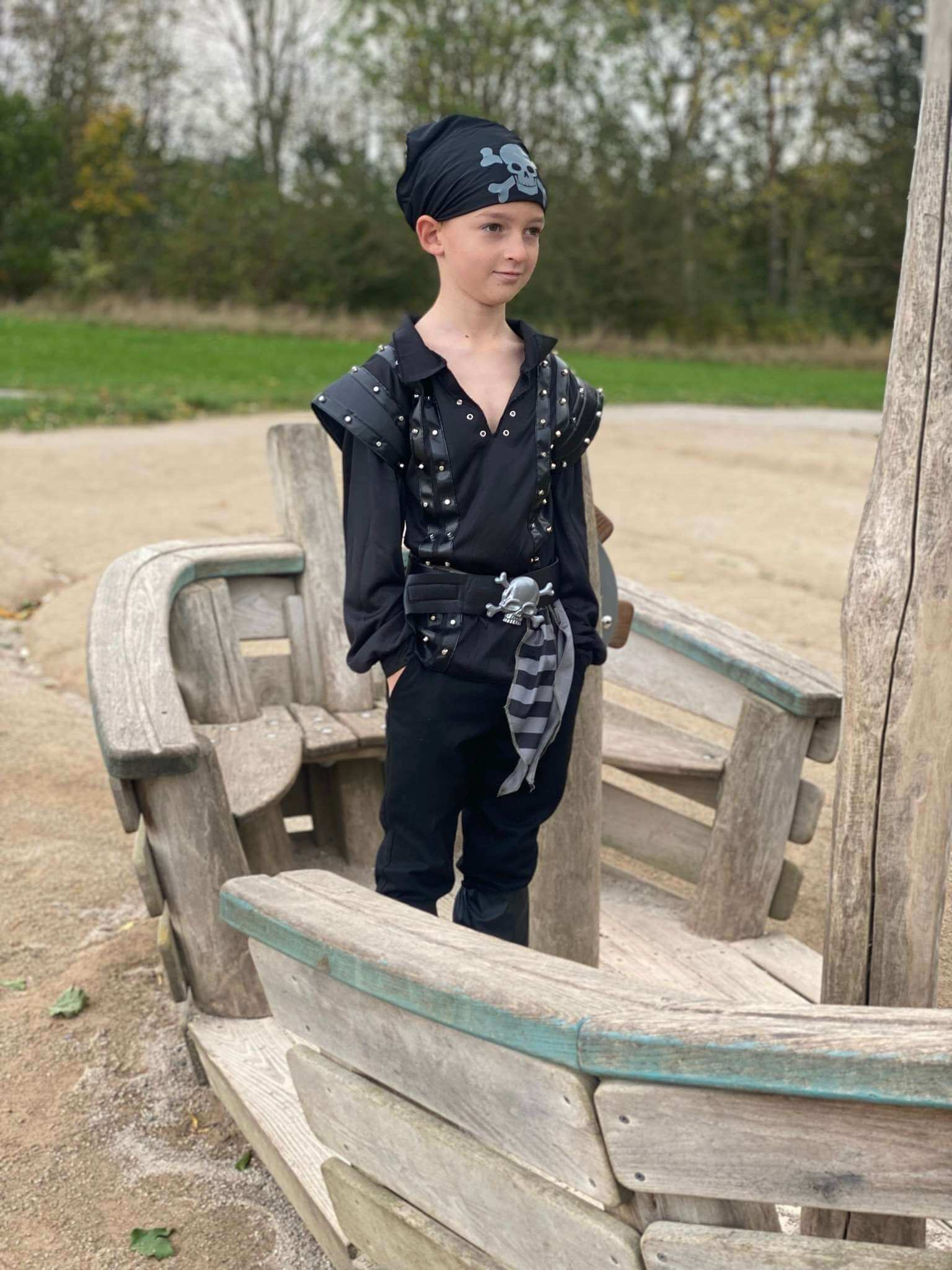 Boy stood on a boat wearing a black pirate costume with studs on the top, a belt, boot covers and a bandana.