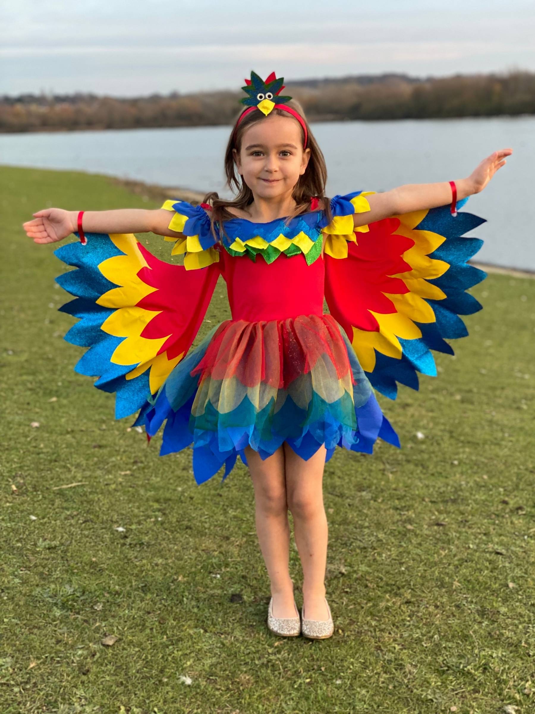 Girl stood on grass in front of water wearing macaw parrot costume.