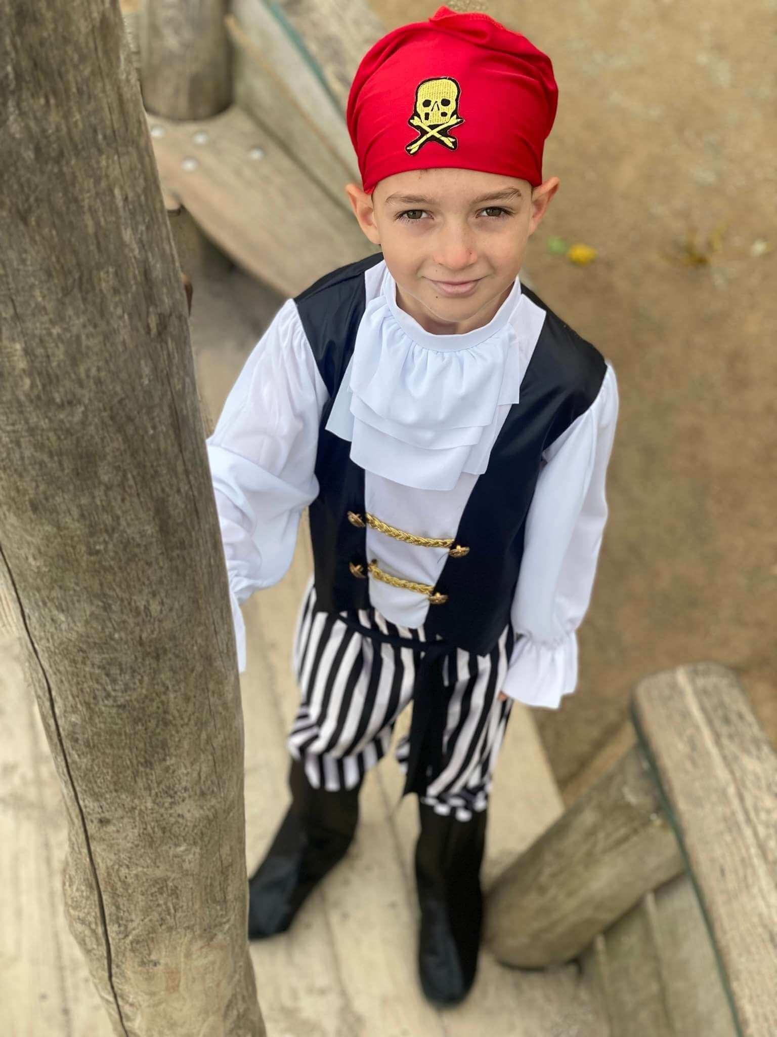 Boy stood on a wooden boat wearing the pirate fancy dress costume showing the skull and crossbones on the bandana. Also shows the detail of the ruffle collar and the golden waistcoat design. 