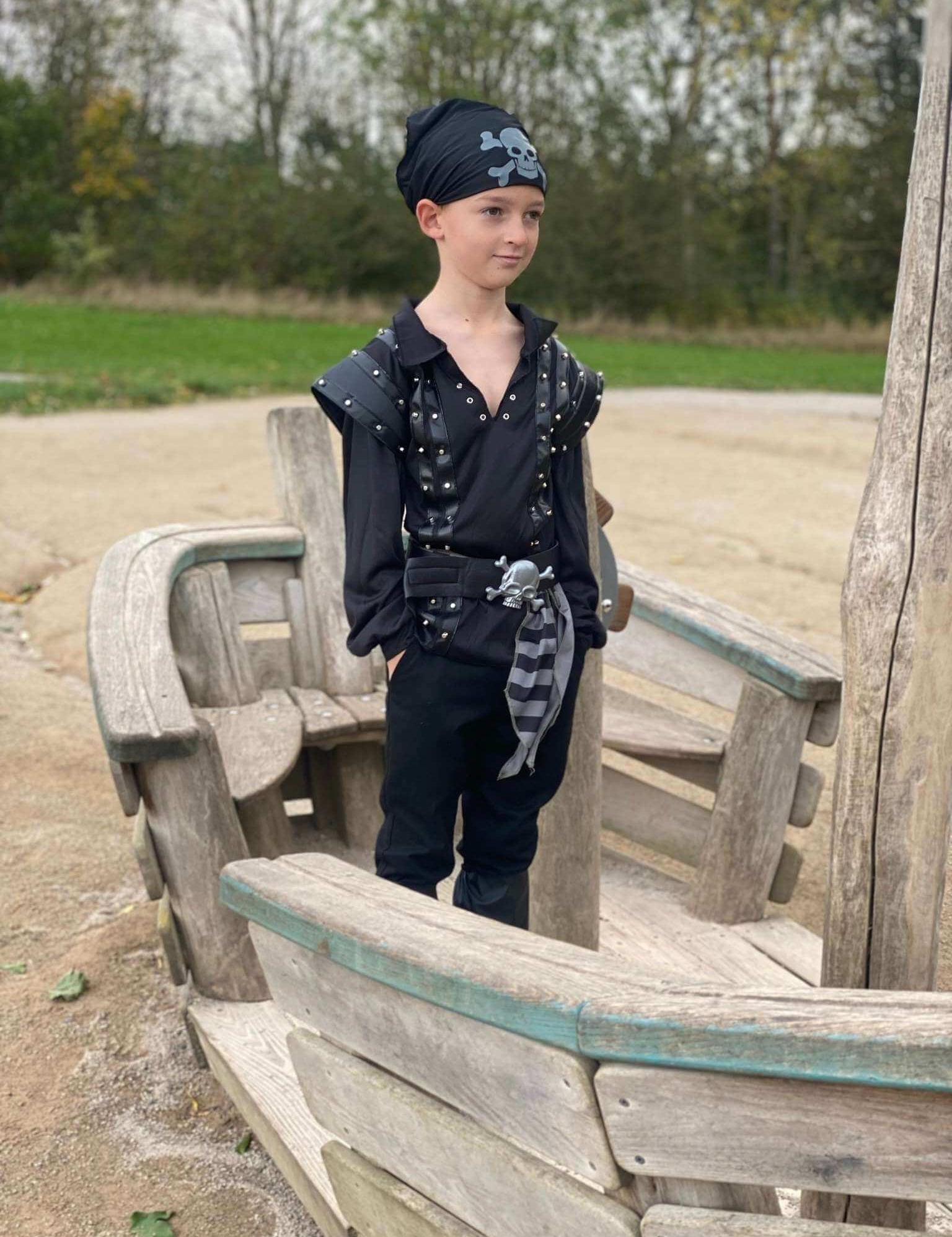 Boy stood on a boat wearing a black pirate costume with studs on the top, a belt, boot covers and a bandana.