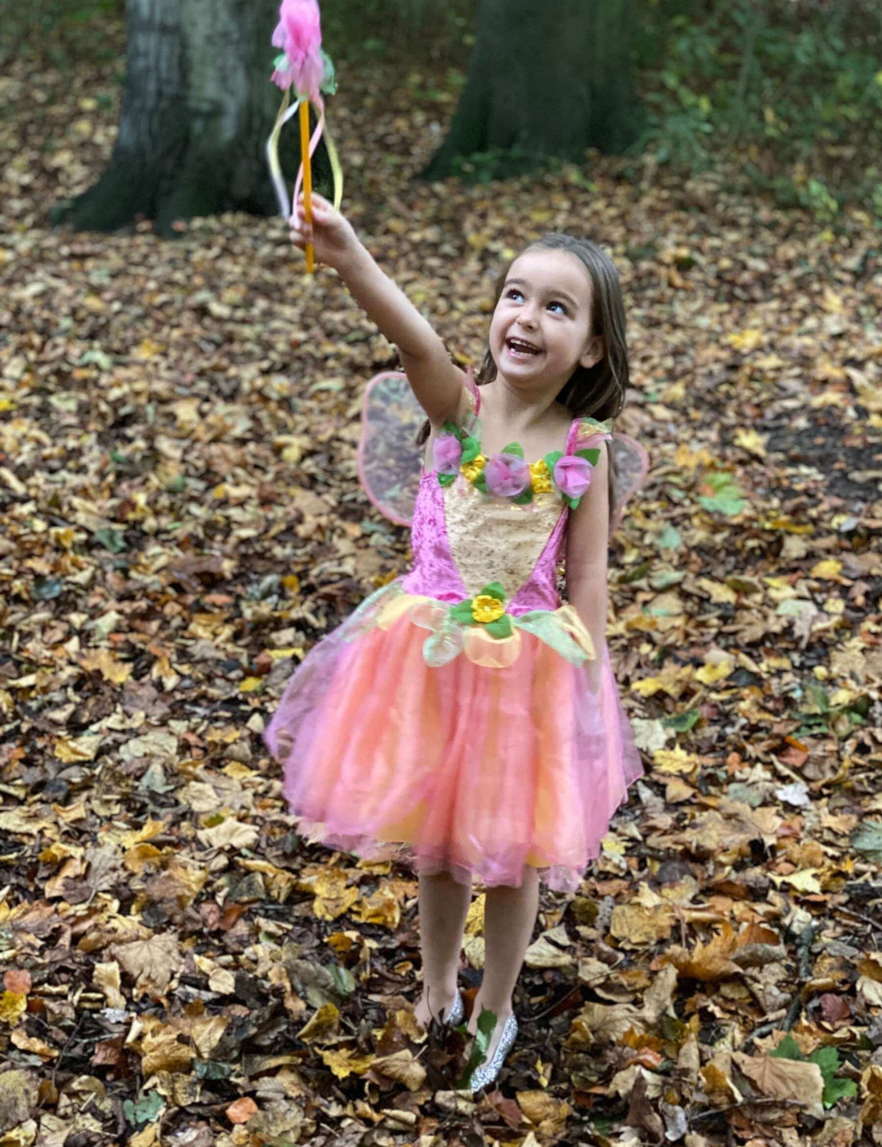 Girl stood in autumn leaves wearing a pink and yellow fairy costume holding a wand in the air.