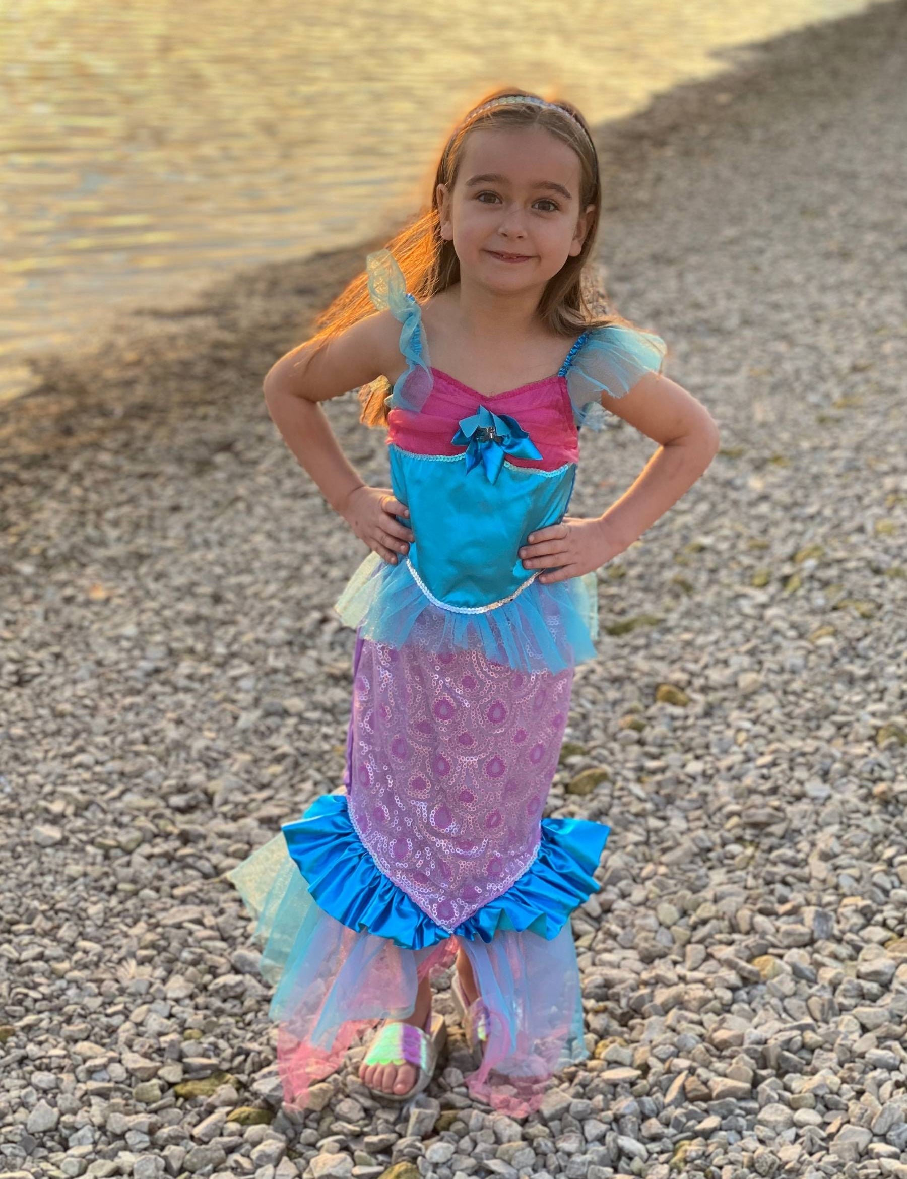 Girl stood next to water with hands on hips wearing blue and pink mermaid dress at sunset.