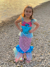 Girl stood next to water with hands on hips wearing blue and pink mermaid dress at sunset.