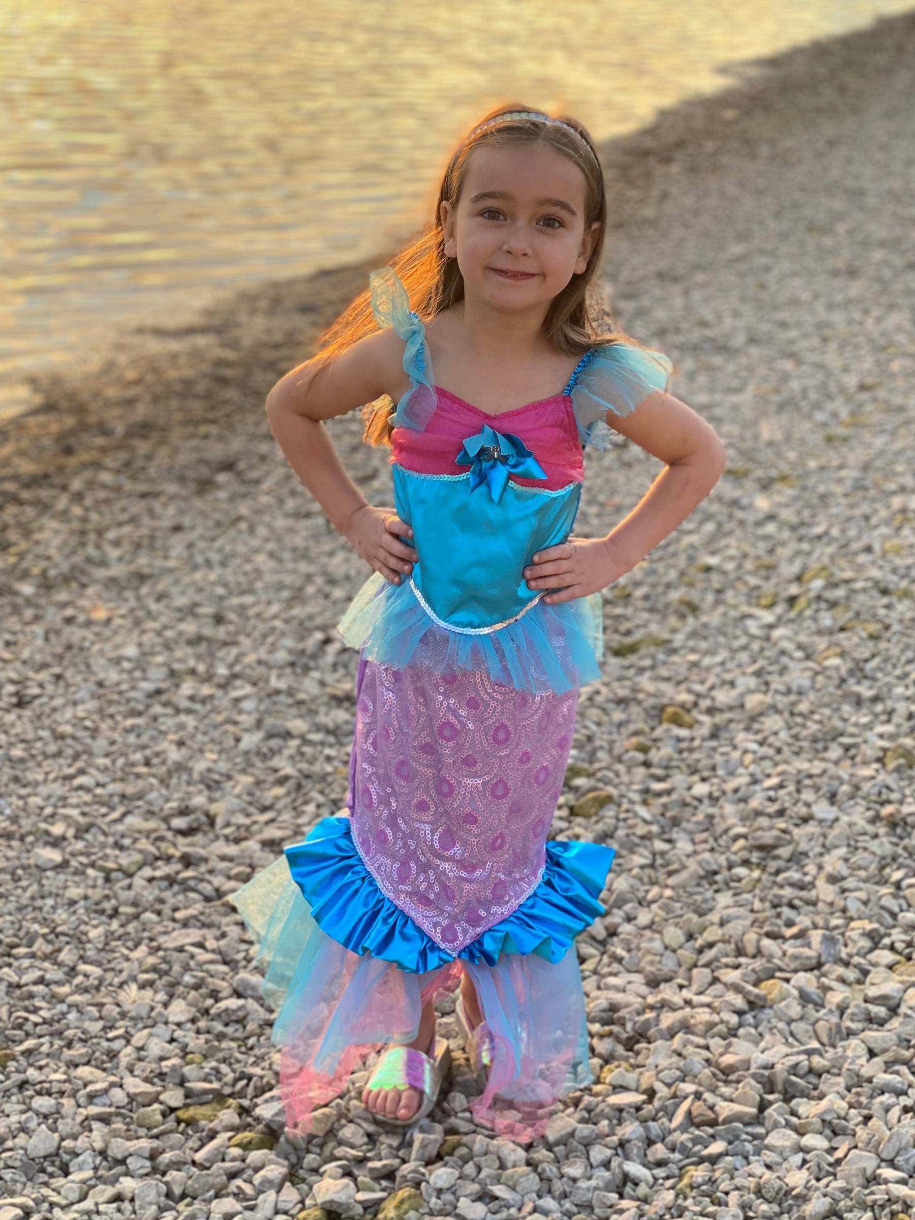 Girl stood next to water with hands on hips wearing blue and pink mermaid dress at sunset.