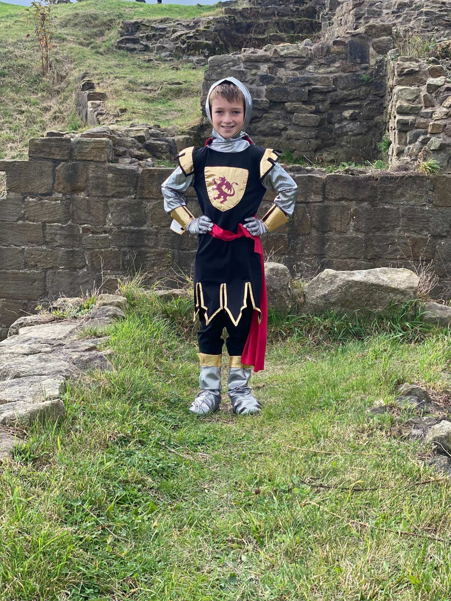 Boy stood in front of castle ruins wearing the Knight costume with his hands on his hips.